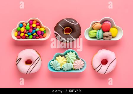 Bonbons et meringues dans un bol en forme de nuage, chocolat avec garniture colorée et donut rose isolé sur fond rose plat Lay Top View Knolling Banque D'Images