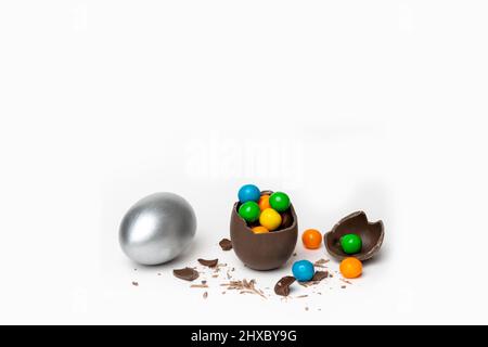 Œuf de pâques au chocolat craqué avec de petites friandises rondes colorées et des chocolats et un œuf d'argent peint entier sur fond blanc, espace de copie. Pâques Banque D'Images