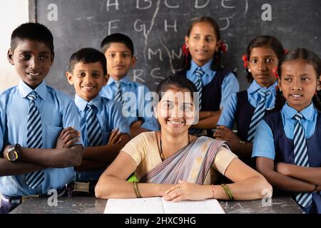 Un bon professeur avec des élèves qui regardent la caméra en salle de classe devant le tableau noir - concept de soutien, d'éducation, de croissance, d'emploi et d'emploi. Banque D'Images