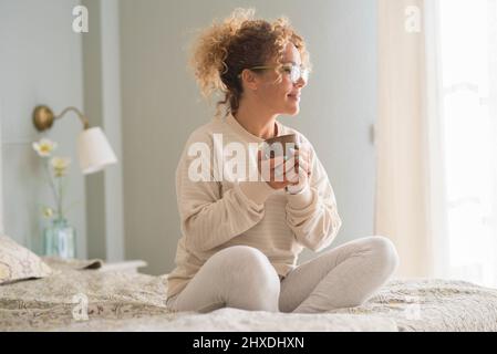 Bonne bonne bonne positive réveil femme adulte assis sur le lit et boire souriant à la lumière de la fenêtre. Les femmes ont un style de vie sain et apprécient le nouveau comi jour Banque D'Images