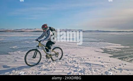 Une femme est à vélo sur la glace. La fille est vêtue d'une veste en duvet argenté, d'un sac à dos de cyclisme et d'un casque. Glace du lac Baïkal gelé. Pneus sur bik Banque D'Images