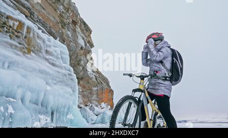 La femme est à vélo près de la grotte de glace. La roche avec des grottes de glace et des glaçons. La fille est vêtue d'une veste en duvet argenté, d'un sac à dos de cyclisme et d'un casque. Banque D'Images