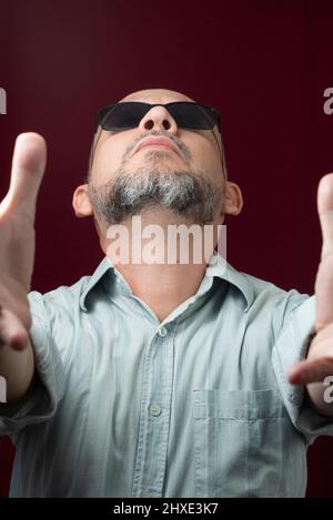 Portrait d'un homme barbu et chauve portant des lunettes sur un fond rouge foncé. Salvador, Bahia, Brésil. Banque D'Images