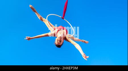 acrobat athlétique, jeune gymnaste gracieuse effectuant l'exercice aérien dans l'air ring à l'extérieur sur fond de ciel. femme flexible en costume rouge se produit Banque D'Images