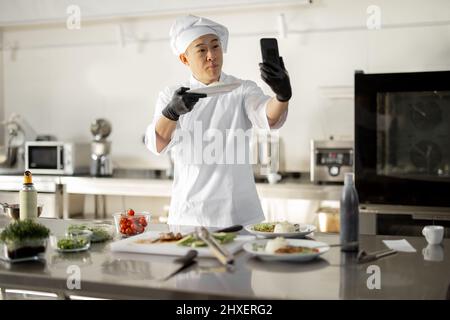 Chef asiatique enregistrant un vlog culinaire au téléphone, montrant une assiette avec repas cuit dans la cuisine. Concept de blog vidéo et de repas de cuisine pour le restaurant Banque D'Images