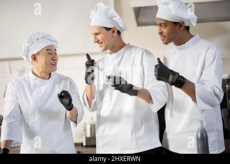 Trois cuisiniers avec différentes spécialités goûtant des plats cuisinés, inventant de nouveaux plats pour un menu. Chef caucasien avec des cuisiniers latins et asiatiques en uniforme travaillant ensemble. Concept de travail d'équipe et cuisine haute Banque D'Images