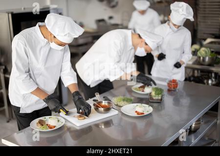 Groupe multiracial de cuisiniers finissant les cours principaux tout en travaillant ensemble dans la cuisine. Cuisiniers portant un uniforme et un masque. L'équipe prépare les repas pour le restaurant Banque D'Images