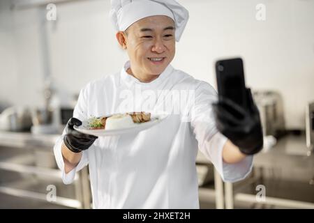 Chef asiatique enregistrant un vlog culinaire au téléphone, montrant une assiette avec repas cuit dans la cuisine. Concept de blog vidéo et de repas de cuisine pour le restaurant Banque D'Images