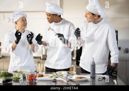 Trois cuisiniers avec différentes spécialités goûtant des plats cuisinés, inventant de nouveaux plats pour un menu. Chef caucasien avec des cuisiniers latins et asiatiques en uniforme travaillant ensemble. Concept de travail d'équipe et cuisine haute Banque D'Images