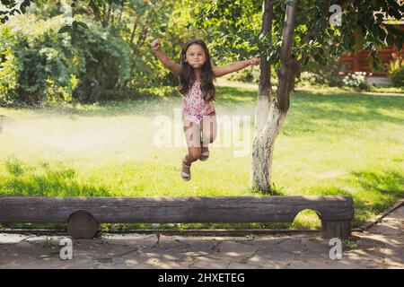 Une petite fille qui brille dans le journal de saut sourire avec des joues bouffies regardant la caméra avec des arroseurs d'eau et un brin vert en arrière-plan dans la journée. Rêver Banque D'Images