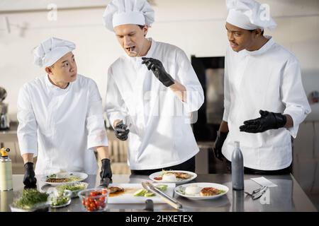 Trois cuisiniers avec différentes spécialités goûtant des plats cuisinés, inventant de nouveaux plats pour un menu. Chef caucasien avec des cuisiniers latins et asiatiques en uniforme travaillant ensemble. Concept de travail d'équipe et cuisine haute Banque D'Images