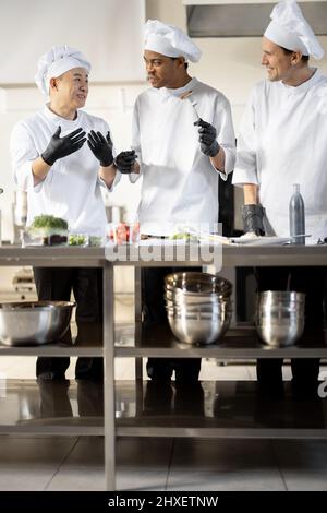 Trois cuisiniers avec différentes spécialités goûtant des plats cuisinés, inventant de nouveaux plats pour un menu. Chef asiatique avec des cuisiniers latino et européens en uniforme travaillant ensemble. Concept de travail d'équipe et cuisine haute Banque D'Images