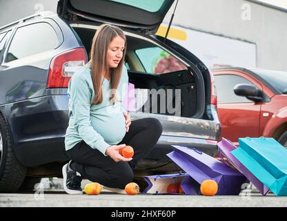 Femme enceinte assise pour ramasser des fruits orange épars du sol contre la voiture avec le coffre ouvert. Femme enceinte ayant commencé des contractions de l'utérus sur le parking après l'épicerie. Banque D'Images