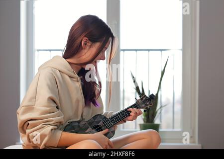 Adolescente avec cheveux violets jouant ukulele guitare dans sa chambre Banque D'Images