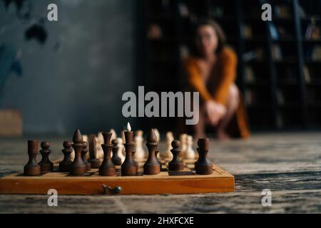 Gros plan sélectif sur le jeu d'échecs sur parquet vintage, femme floue et méconnaissable assise loin près des étagères de la bibliothèque sombre. Banque D'Images