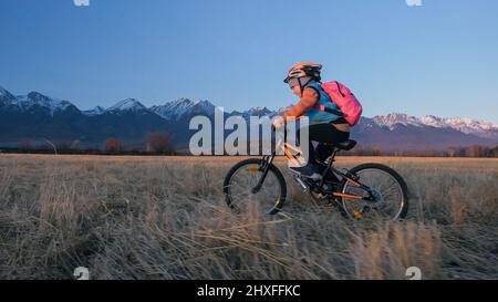 L'un des enfants de race blanche rides vélo en champ de blé. Little girl riding noir orange cycle sur fond de belles montagnes enneigées. Motion Biker ride Banque D'Images