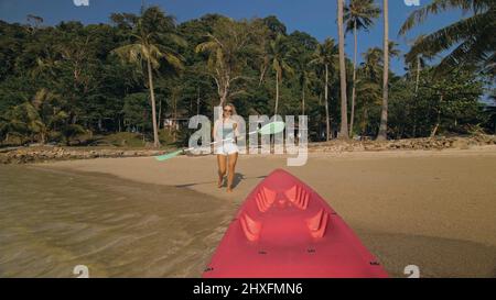 Jeune femme et lunettes de soleil tire le canot en plastique rose avec la pagaie pour azurer l'eau de mer de la plage avec les palmiers. Début de la sortie en bateau, embarquement. Tr Banque D'Images