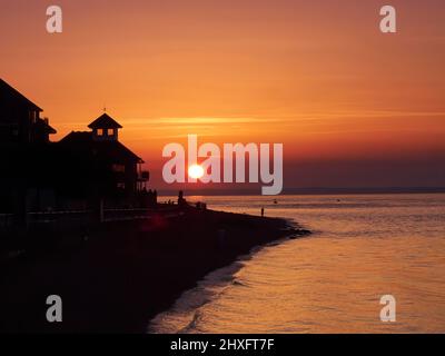 Un coucher de soleil richement coloré vu de Copes Beach, la lumière du soleil venant par les fenêtres d'un bâtiment de bord de mer et silhoueted des gens sur la plage. Banque D'Images