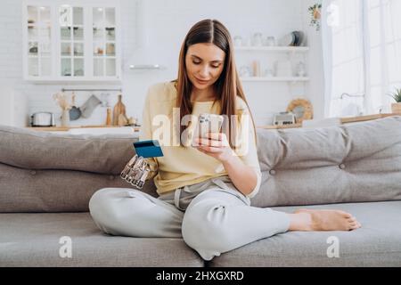 Jolie femme heureuse avec bras de prothèse paie pour la commande en ligne avec carte de crédit via téléphone mobile sur le canapé dans la salle de séjour à proximité Banque D'Images