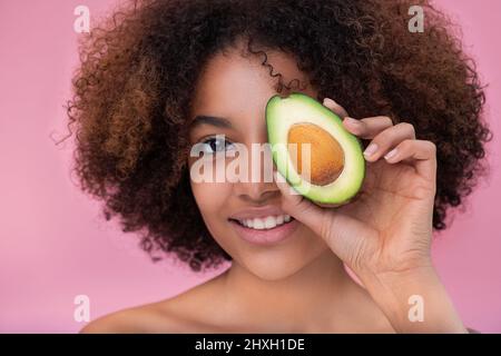 Portrait d'une belle jeune femme à la peau sombre avec des cheveux bouclés couvre un oeil avec la moitié d'un avocat regarde l'appareil photo et sourit sur un fond rose. Banque D'Images