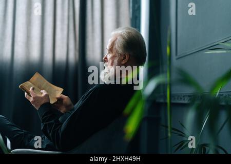 Vue latérale d'un homme âgé aux cheveux gris concentré lisant un livre à la maison assis à table, détendez-vous et reposez-vous avec de la littérature. Banque D'Images