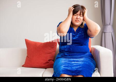 Une femme asiatique de l'embonpoint est assise sur le canapé dans le salon. Et tenir la main dans la tête à cause de maux de tête de migraine. Concepts de illnes Banque D'Images