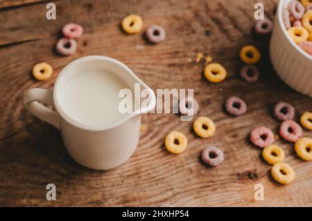 Le lait dans un pot à lait et les flocons de fruits ronds colorés dans un bol blanc sont dispersés sur une table en bois marron Banque D'Images