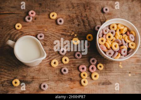 Le lait dans un pot à lait et les flocons de fruits ronds colorés dans un bol blanc sont dispersés sur une table en bois marron Banque D'Images