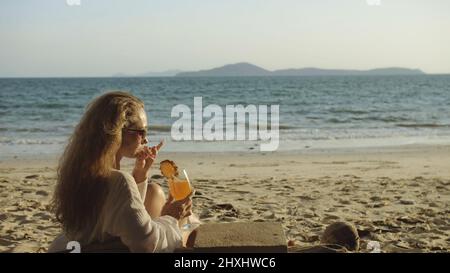 Belle femme assise dans une robe blanche lunettes de soleil, boire un cocktail d'ananas Pina Colada.Belle plage, vagues au coucher du soleil.Concept repos tropique Banque D'Images