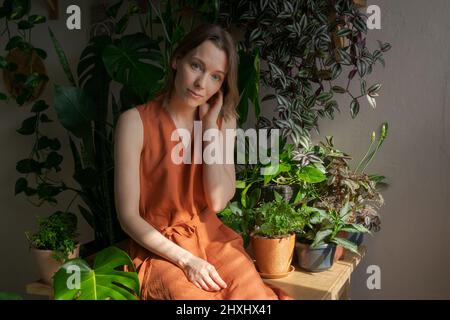 Femme jardinière à la maison, soin des plantes à la maison, verdure à la maison, amour des plantes, jardin intérieur confortable. Une fille caucasienne dans une robe orange est assise sur une table et tient un pot de fleur Banque D'Images