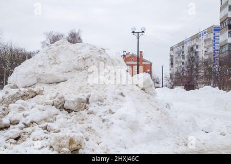 Une puissante dérive de neige au bord de la route sur fond de maisons de ville et d'arbres. Sur la route se trouve de la neige sale dans des tas hauts. Paysage urbain d'hiver. Jour d'hiver nuageux, lumière douce. Banque D'Images