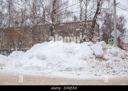 Une énorme dérive de neige au bord de la route sur fond de maisons de ville et d'arbres. Sur la route se trouve de la neige sale dans des tas hauts. Paysage urbain d'hiver. Jour d'hiver nuageux, lumière douce. Banque D'Images