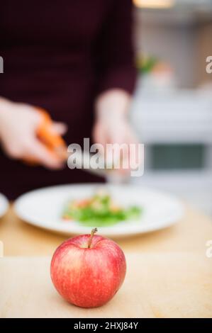 Pomme rouge sur la table. Préparation de salade en arrière-plan. Concentrez-vous sur la pomme, très faible profondeur de champ. Banque D'Images