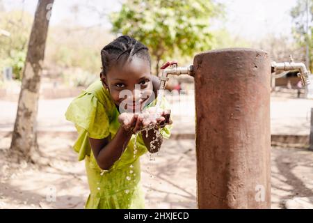 Gaie fille africaine noire dans une jolie robe jaune de boire de l'eau douce d'un robinet public Banque D'Images