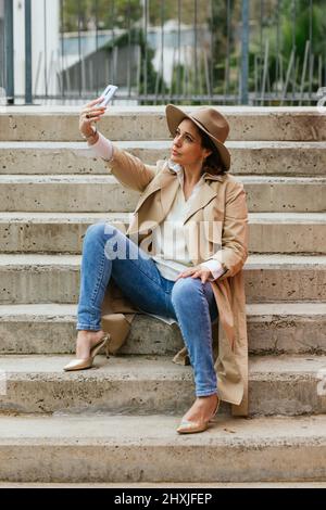 Femme vêtue de vêtements chauds et élégants et emportant un selfie avec un téléphone portable tout en étant assise dans les escaliers de la rue. Banque D'Images