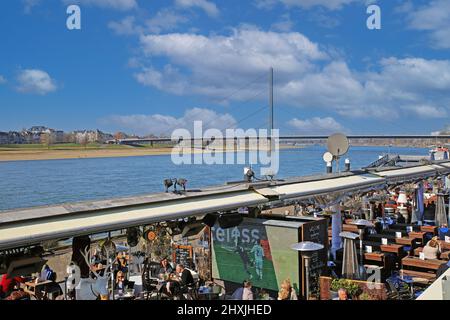 Düsseldorf (Rheinpromenade), Allemagne - mars 9. 2022: Vue sur le café-restaurant extérieur avec des personnes assises le jour d'hiver ensoleillé, le Rhin et Oberkas Banque D'Images