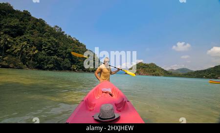 Jeune homme en chapeau et lunettes de soleil tire à l'aide de la corde de plastique rose canoë avec pagaie pour azurer l'eau de mer de la plage avec des palmiers. Voyage dans un pays tropical Banque D'Images