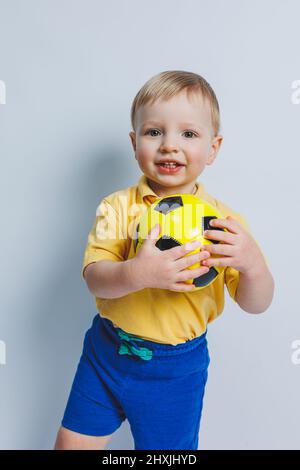 Un petit garçon avec un ballon de football dans ses mains sur fond blanc, un enfant est un joueur de football débutant, un sport pour les enfants. Petit athlète. Jaune Banque D'Images