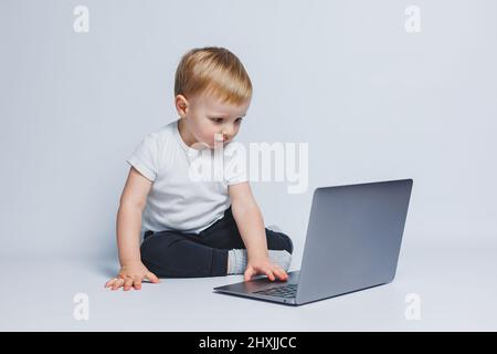 Un petit garçon de 3-4 ans est assis avec un ordinateur portable sur un fond blanc. Un enfant dans un T-shirt blanc et un pantalon noir regarde un ordinateur portable. Enfants modernes Banque D'Images