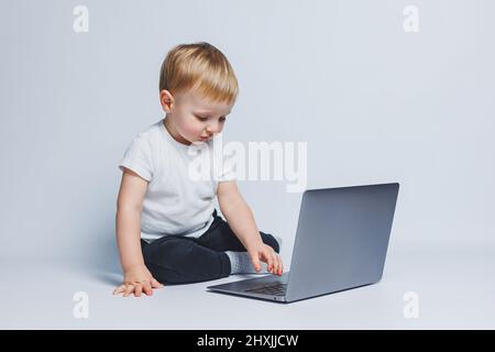 Un petit garçon de 3-4 ans est assis avec un ordinateur portable sur un fond blanc. Un enfant dans un T-shirt blanc et un pantalon noir regarde un ordinateur portable. Enfants modernes Banque D'Images