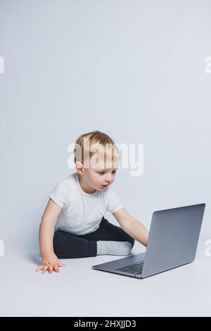 Un petit garçon de 3-4 ans est assis avec un ordinateur portable sur un fond blanc. Un enfant dans un T-shirt blanc et un pantalon noir regarde un ordinateur portable. Enfants modernes Banque D'Images