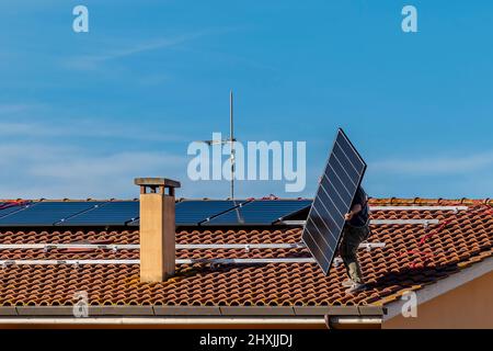 Un technicien survolant un toit en tuiles rouges soulève un panneau solaire qu'il est sur le point d'ajouter à un système photovoltaïque en cours d'installation Banque D'Images