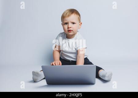 Un jeune garçon de 3-4 ans est assis avec un ordinateur portable sur un fond blanc. Un enfant dans un T-shirt blanc et un pantalon noir est assis à un ordinateur portable et regarde le CA Banque D'Images