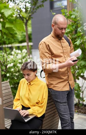 Équipe d'affaires travaillant à l'extérieur du parc. Femme d'affaires gaie assise au travail avec un ordinateur portable tandis que l'homme d'affaires utilise un téléphone cellulaire. Les gens d'affaires de la ville. Image de haute qualité Banque D'Images