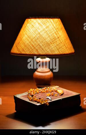 Lumière douce de la lampe de table, vieux livre (livre de prière ou Bible) et rosaire catholique ambre sur la table. Religion, prière du soir. Religion, heure de la soirée prier Banque D'Images