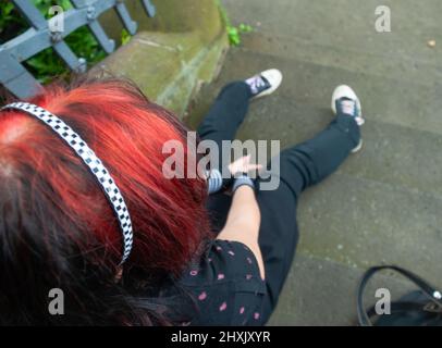 Punk emo girl, jeune adulte avec des cheveux rouges noirs, assis sur un escalier extérieur, horizontal Banque D'Images