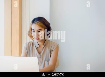 Femme asiatique, freelance avec cheveux courts tendance regardant l'écran tout en travaillant avec ordinateur portable sur fond blanc mur avec espace de copie, à proximité Banque D'Images