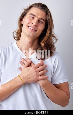 Joyeux sourire caucasien gars exprimant la gratitude, reconnaissant pour tout, garder les mains sur la poitrine et regarder la caméra, isolé sur le dos gris clair Banque D'Images