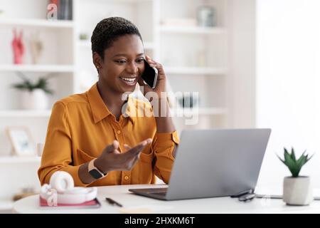 Femme souriante assise à la table avec un ordinateur portable, ayant une conversation téléphonique Banque D'Images