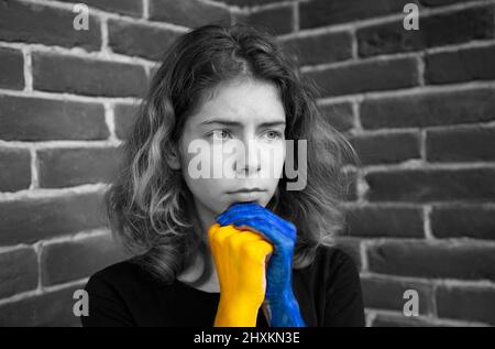 Noir - Portrait blanc d'une jeune femme triste, mains peintes en couleurs de drapeau ukrainien. Accent de couleur sur le jaune et le bleu. Arrêter la guerre. Espoir et foi. Banque D'Images